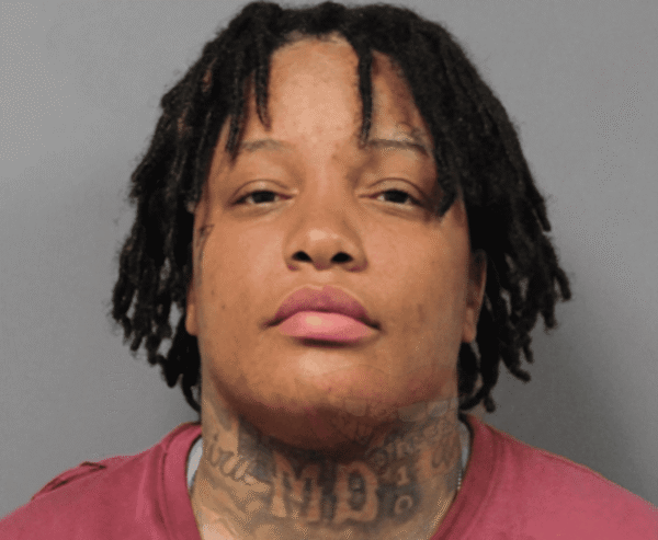 Mugshot of a young man with medium-length dreadlocks, wearing a pink shirt, against a gray background; tattoos visible on his neck.
