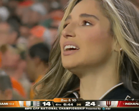 Close-up of a smiling woman with long blonde hair reacting at a college football game, blurred crowd in the background.