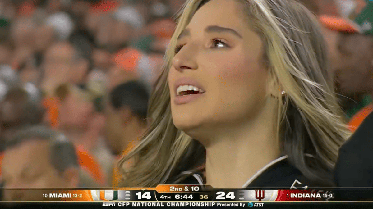 Close-up of a smiling woman with long blonde hair reacting at a college football game, blurred crowd in the background.