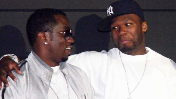 Two men posing and smiling together, one with sunglasses and silver chains, the other wearing a tilted baseball cap and a white shirt with a chain, arm around the other.