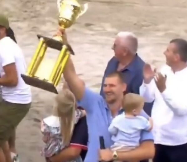 Man in a blue shirt raises a large gold trophy in celebration while a crowd cheers nearby, on a sandy outdoor area.