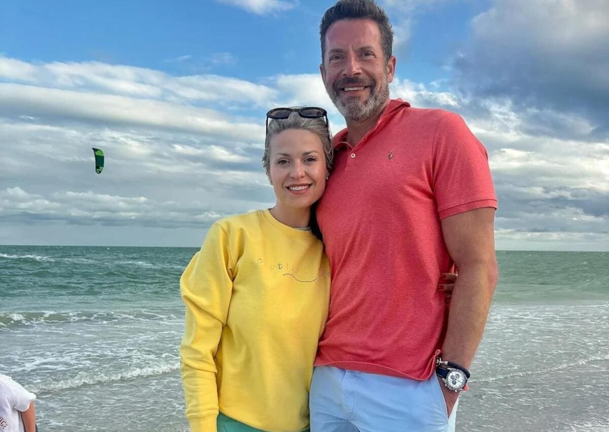 Smiling couple stands on a beach, the woman in a yellow sweatshirt and sunglasses on her head, the man in a coral polo and light shorts, with ocean waves behind them and a kite surfer in the distance.