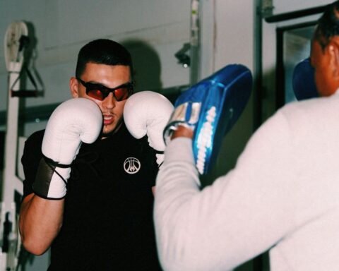 Boxer in white gloves and sunglasses facing a coach holding a blue focus pad in a gym.",