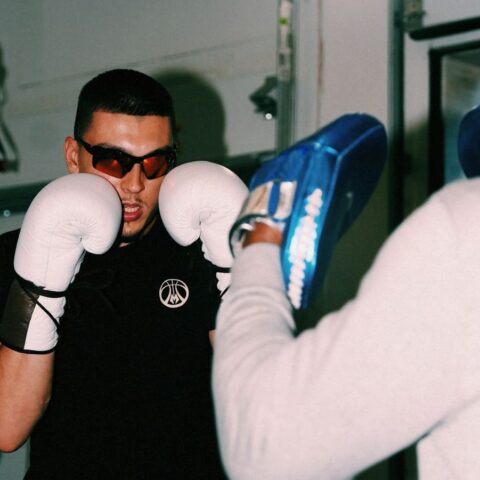 Boxer in white gloves and sunglasses facing a coach holding a blue focus pad in a gym.",