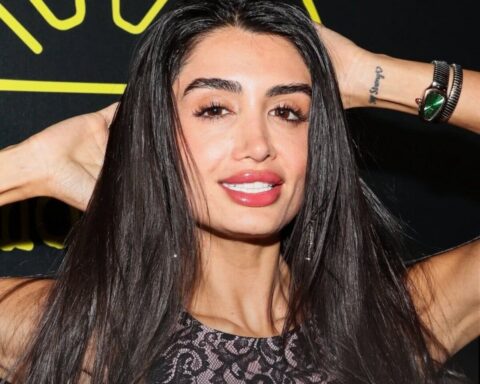 Woman with long dark hair posing with hands behind her head, wearing a black lace top and a silver bracelet with a green stone watch.