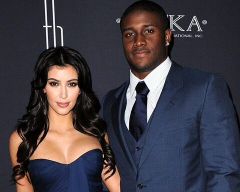 Woman with long dark hair in a navy strapless gown posing next to a tall man in a blue suit at a formal event against a dark backdrop with logos.