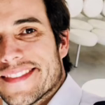 Close-up of a smiling man in a white shirt taking a selfie, with white cushioned chairs in the bright background.