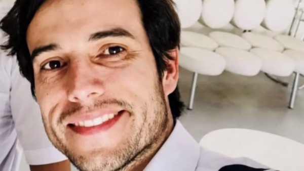 Close-up of a smiling man in a white shirt taking a selfie, with white cushioned chairs in the bright background.
