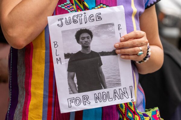 Person holding a protest flyer with a black-and-white portrait and the words 'JUSTICE FOR NOLAN!' and 'BLM' on the sides.