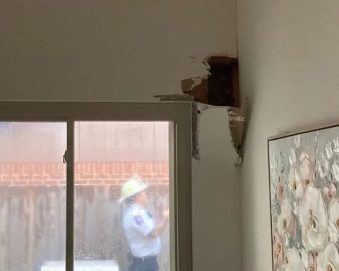 Interior shows a wall with a large hole and torn plaster above a window; a person in a hat is outside viewing the damage through the glass.