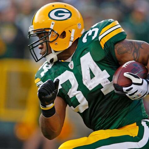 Green Bay Packers running back in green and gold grips the football and sprints on the field during a game, helmet visible.
