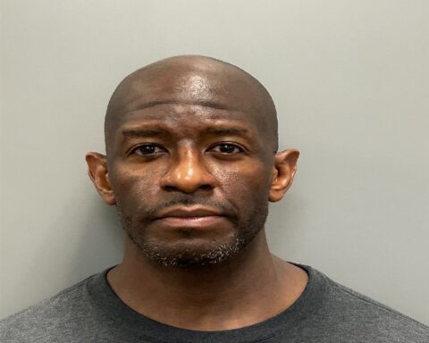 Mugshot of a bald man with a short beard, wearing a dark gray T‑shirt against a plain gray wall.