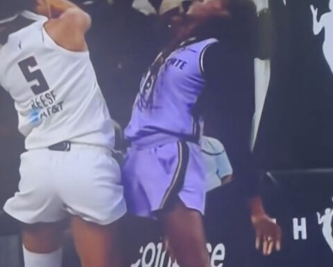 Basketball players in white (number 5) and purple uniforms collide at the sideline during a game near advertising boards.