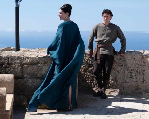Two people stand at a stone railing overlooking the sea: a woman in a flowing teal cloak facing away, and a man in a gray tunic with a belt.