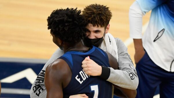 Two basketball players hug on the court during a game or warm‑up, one in a blue jersey with the number 7 and the other in a gray sweatshirt, both facing each other.