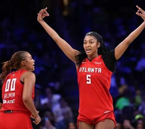 Two female basketball players in red uniforms on a court; one has arms raised in celebration as the other looks on.