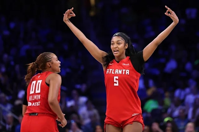 Two female basketball players in red uniforms on a court; one has arms raised in celebration as the other looks on.