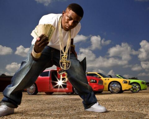 Young man crouching and flashing a stack of cash in front of a row of bright customized cars in an open lot under a blue sky.",