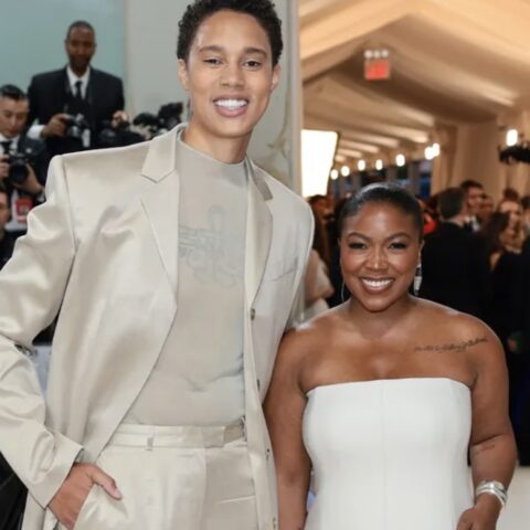 Smiling couple posing on a red carpet with photographers in the background.