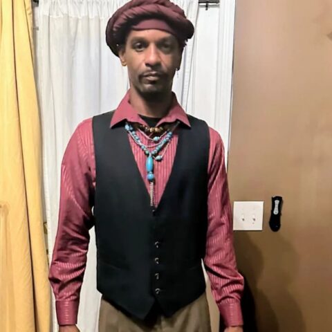 Man indoors wearing a red striped shirt, black vest, tan pants, maroon turban, and turquoise bead necklace as he stands facing the camera.