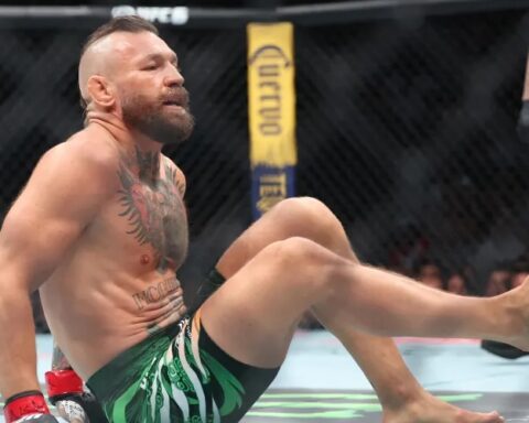 MMA fighter sits on the mat inside the octagon, legs raised, wearing green shorts and red gloves, sweaty and looking forward.