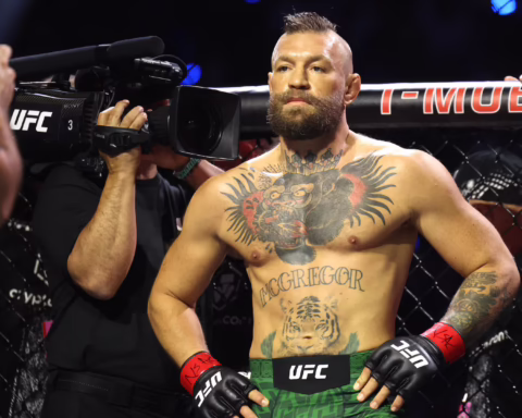 Tattooed UFC fighter in green shorts and red gloves stands in the cage while a cameraman films him from the left.