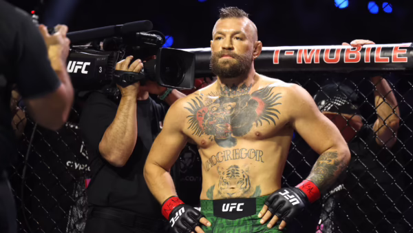 Tattooed UFC fighter in green shorts and red gloves stands in the cage while a cameraman films him from the left.