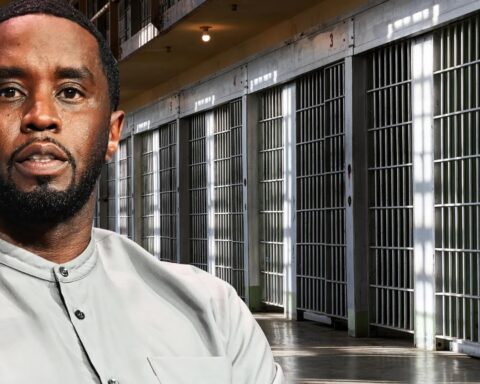 Man in a light gray shirt stands in a prison hallway with rows of barred cells behind him.
