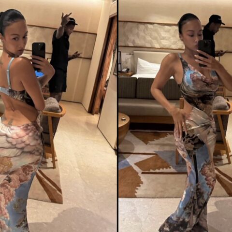 Woman posing in a long, blue floral dress in a hotel room mirror; man in the background making a peace sign.