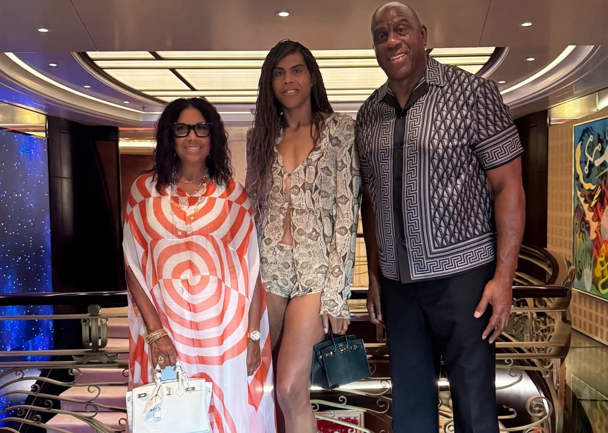 Three adults pose together on a railing indoors; left woman in orange-white swirled dress with white bag, middle woman in snakeskin-print outfit with shorts and green handbag, man on right in geometric patterned shirt and black pants.