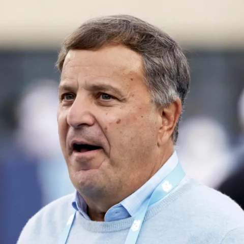 Close-up of a middle-aged man speaking, wearing a light blue sweater and a lanyard at an event