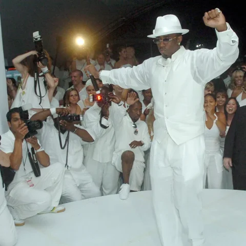 Man in a white suit and fedora dances on a white circular stage as photographers in white attire photograph him from the crowd.