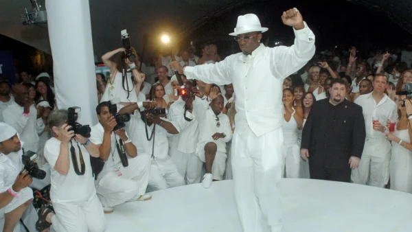 Man in a white suit and fedora dances on a white circular stage as photographers in white attire photograph him from the crowd.