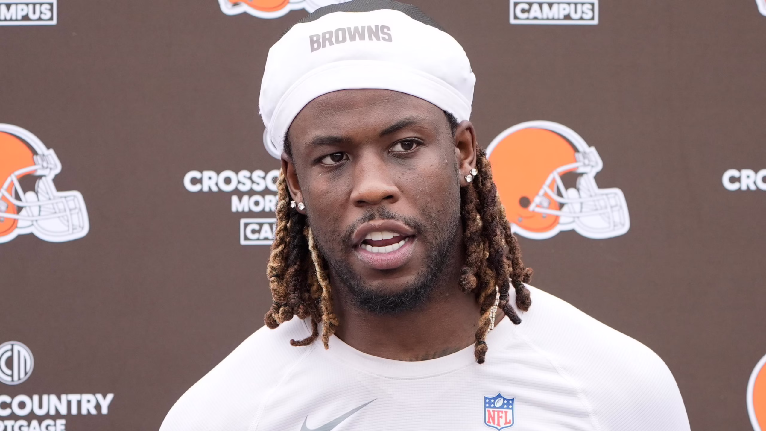 Close-up of a Black NFL player wearing a white Browns headband at a backdrop featuring Browns logos and signage, mid-speech.