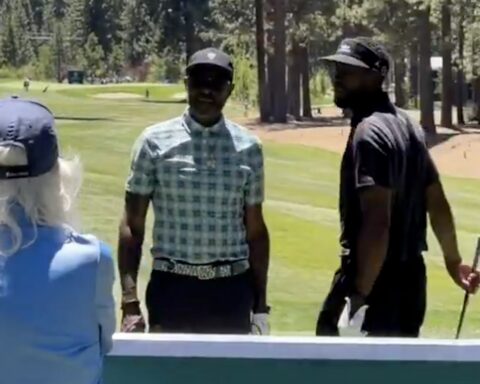 Two men stand on a sunny golf course, one in a checkered shirt and cap, the other in a dark shirt and visor, with trees in the background.