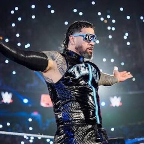 WWE wrestler in a black shiny suit with blue sunglasses raises arms in the ring, lights sparkling around him.