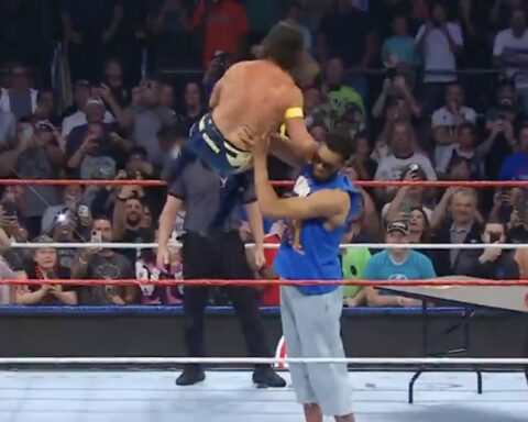 Wrestling match in a crowded arena; masked wrestler in blue pants stands left while a muscular opponent is lifted overhead by a referee or teammate in the center of the ring as spectators film.