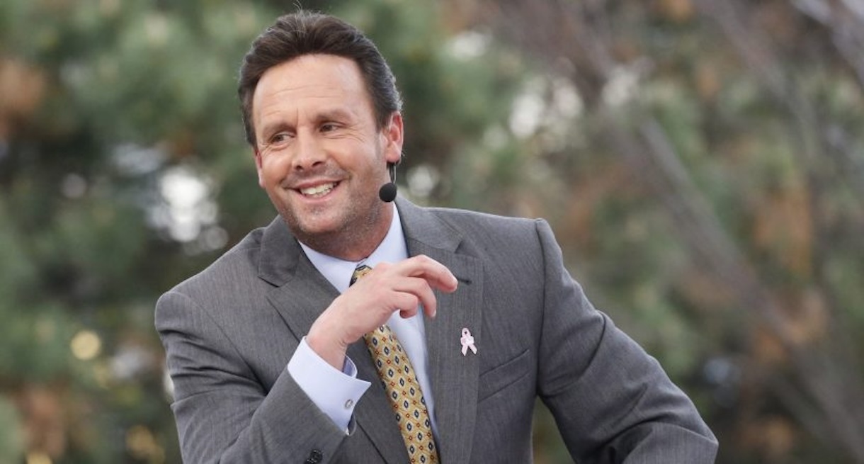 Male public speaker in a gray suit with a pink ribbon pin, wearing a headset mic outdoors and smiling while speaking.