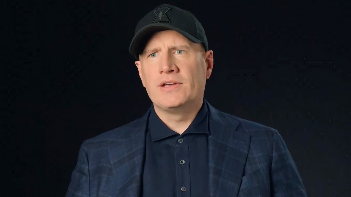 Man in a navy plaid blazer and dark shirt speaks against a solid dark background, wearing a black cap.