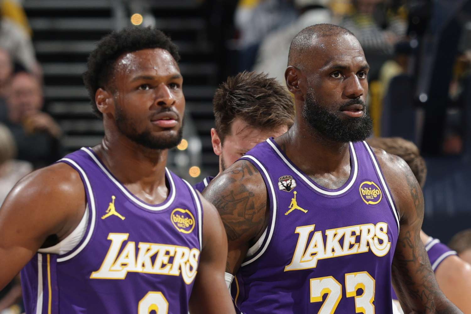 Two Los Angeles Lakers players in purple jerseys stand on the basketball court during a game, focused expressions, blurred crowd in the background.