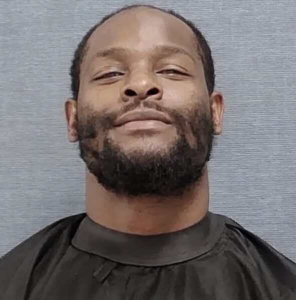 Mugshot of a smiling Black man with a full beard and short hair, wearing a dark shirt against a gray backdrop.