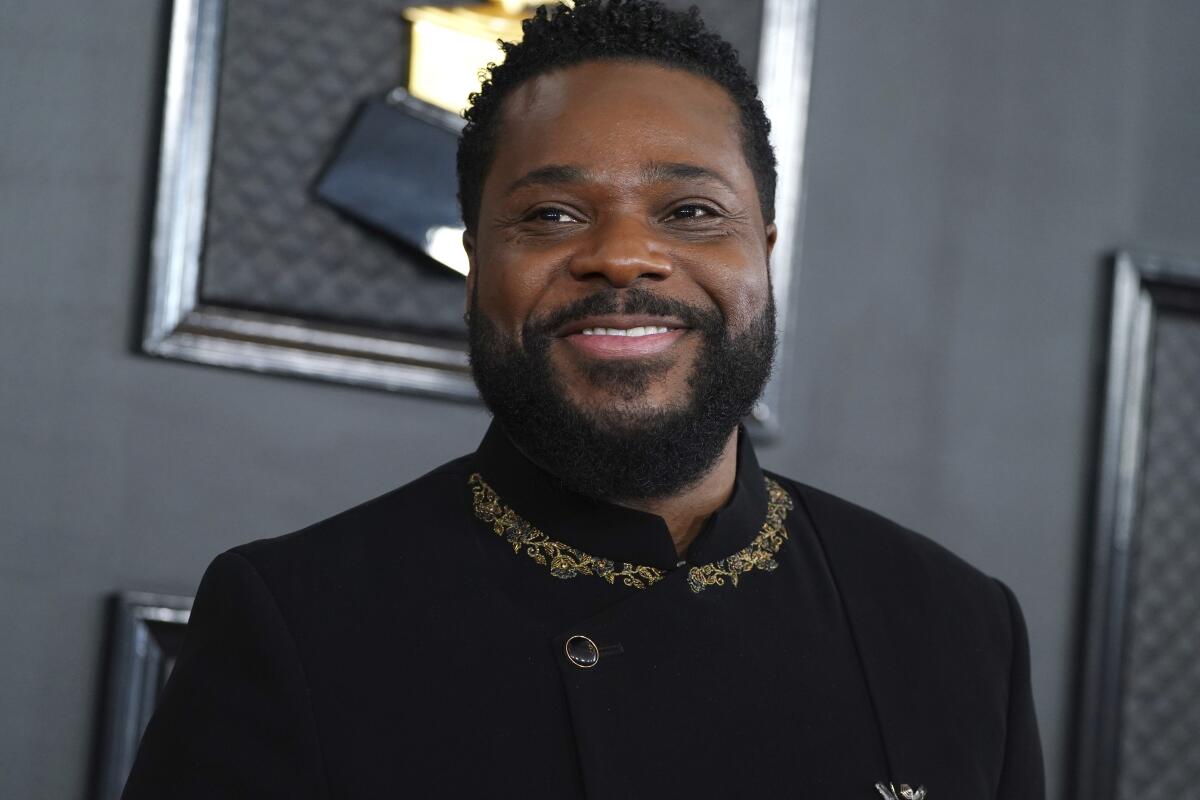 Smiling man with a beard wearing a black suit with gold embroidery at a formal event