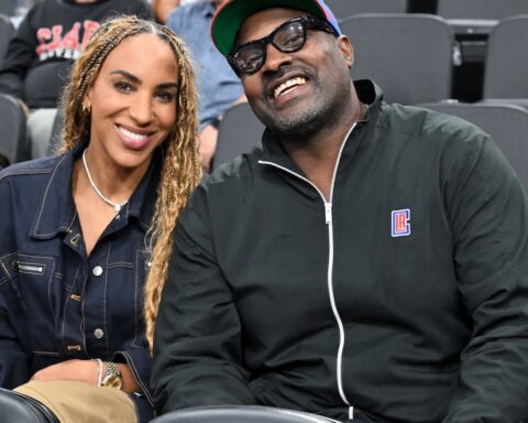 Smiling couple seated in stadium seats; woman with braided hair in a denim jacket, man in a black zip jacket and cap.