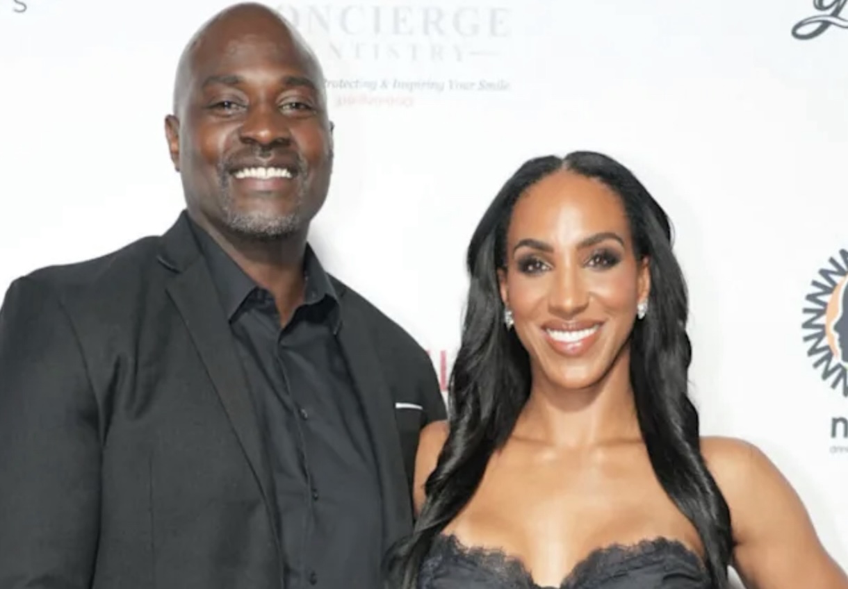 Smiling man in a black suit beside a smiling woman in a black lace dress at a formal event on a white backdrop