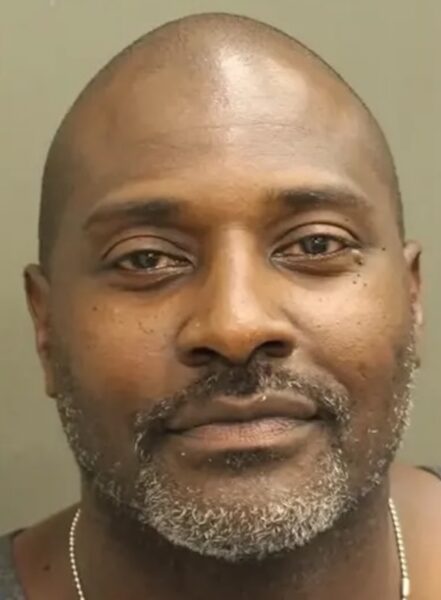 Close-up of a smiling Black man with a gray beard, bald head, and a thin necklace against a neutral background.