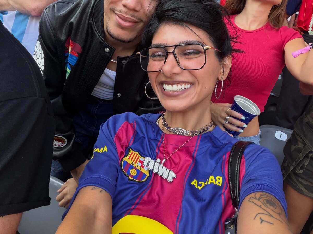 Smiling woman in FC Barcelona jersey and large glasses takes a selfie with friends at a stadium, pink and blue jersey visible.