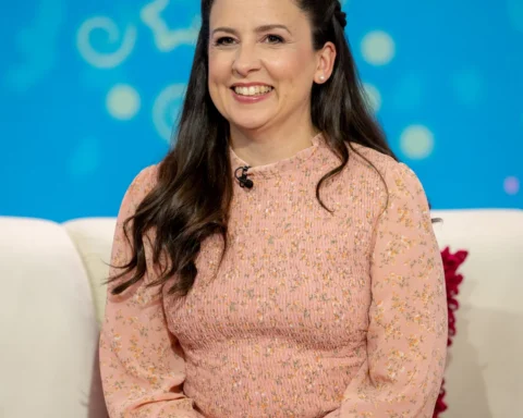 Smiling woman with long dark hair in a pink floral dress seated on a white sofa against a blue studio backdrop with abstract circular patterns.