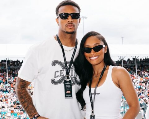 Two smiling people pose together at an outdoor event, wearing sunglasses and lanyards, with a large crowd in the background.