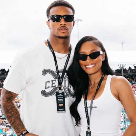 Two smiling people pose together at an outdoor event, wearing sunglasses and lanyards, with a large crowd in the background.