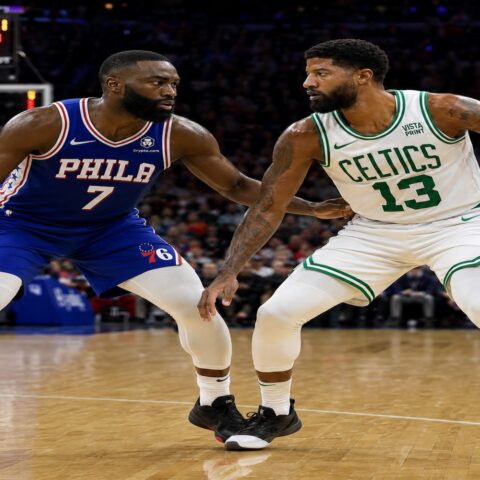 Two basketball players, one in a blue Phillies jersey (number 7) and the other in a white Celtics jersey (number 13), guard each other on a hardwood court during a game, with a scoreboard visible in the background.